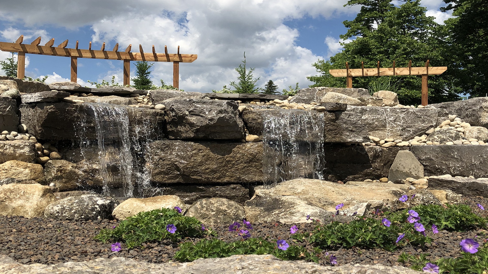 Stone waterfall cascades gently, surrounded by vibrant purple flowers and greenery under a partly cloudy sky, with wooden trellis structures in the background.