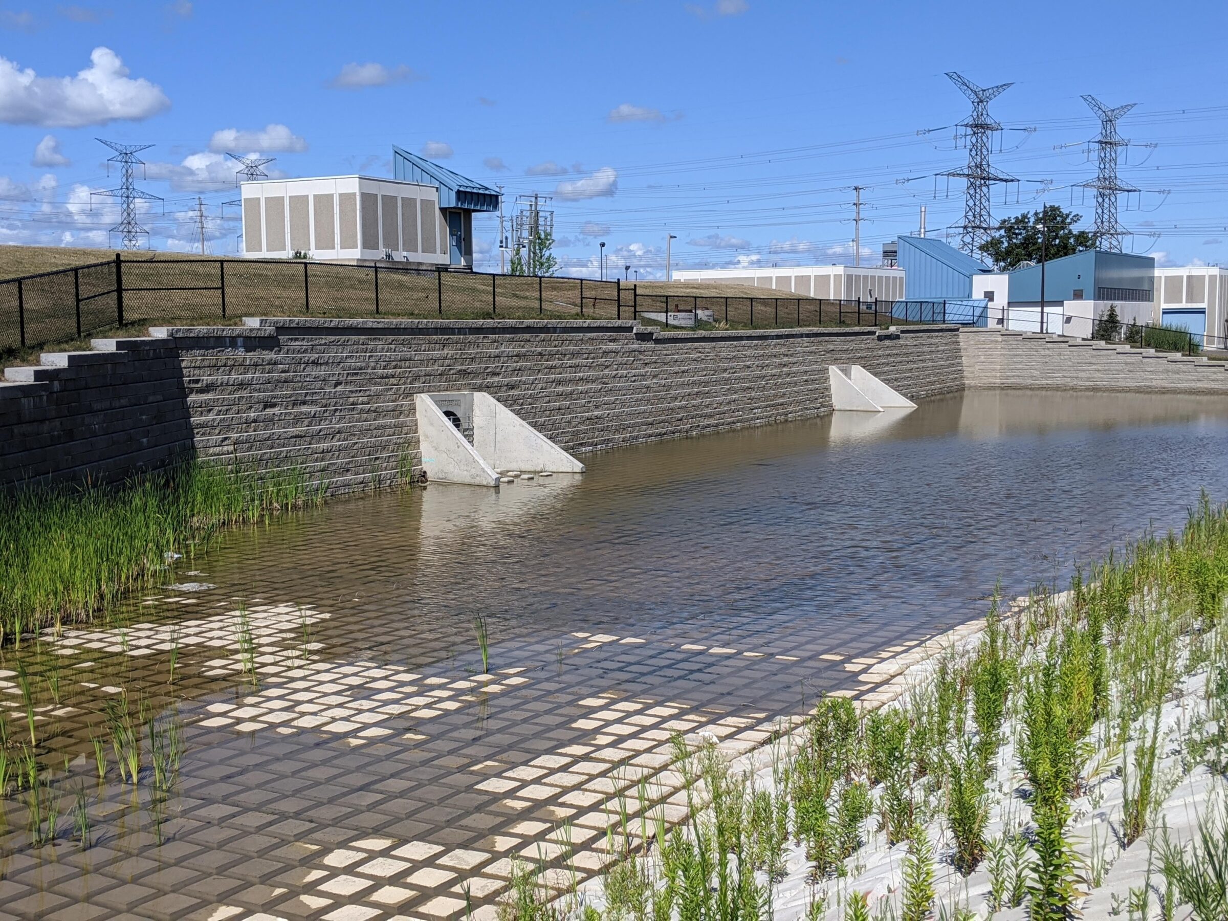 The image shows a water retention area with sloped walls, surrounded by industrial buildings and power lines under a blue sky.