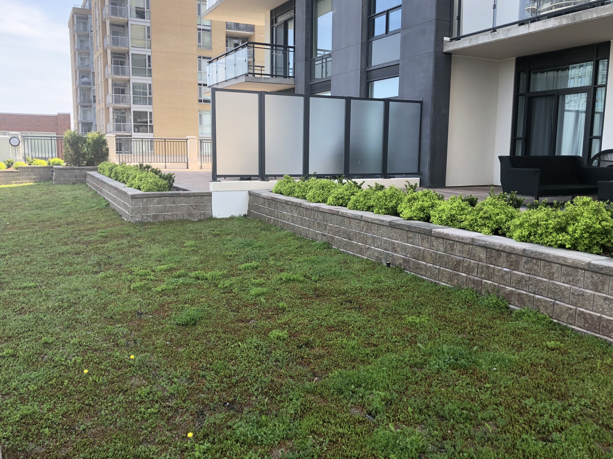 A modern building with glass balconies overlooks a landscaped patio featuring manicured greenery. Cozy outdoor furniture is present, enhancing the tranquil setting.