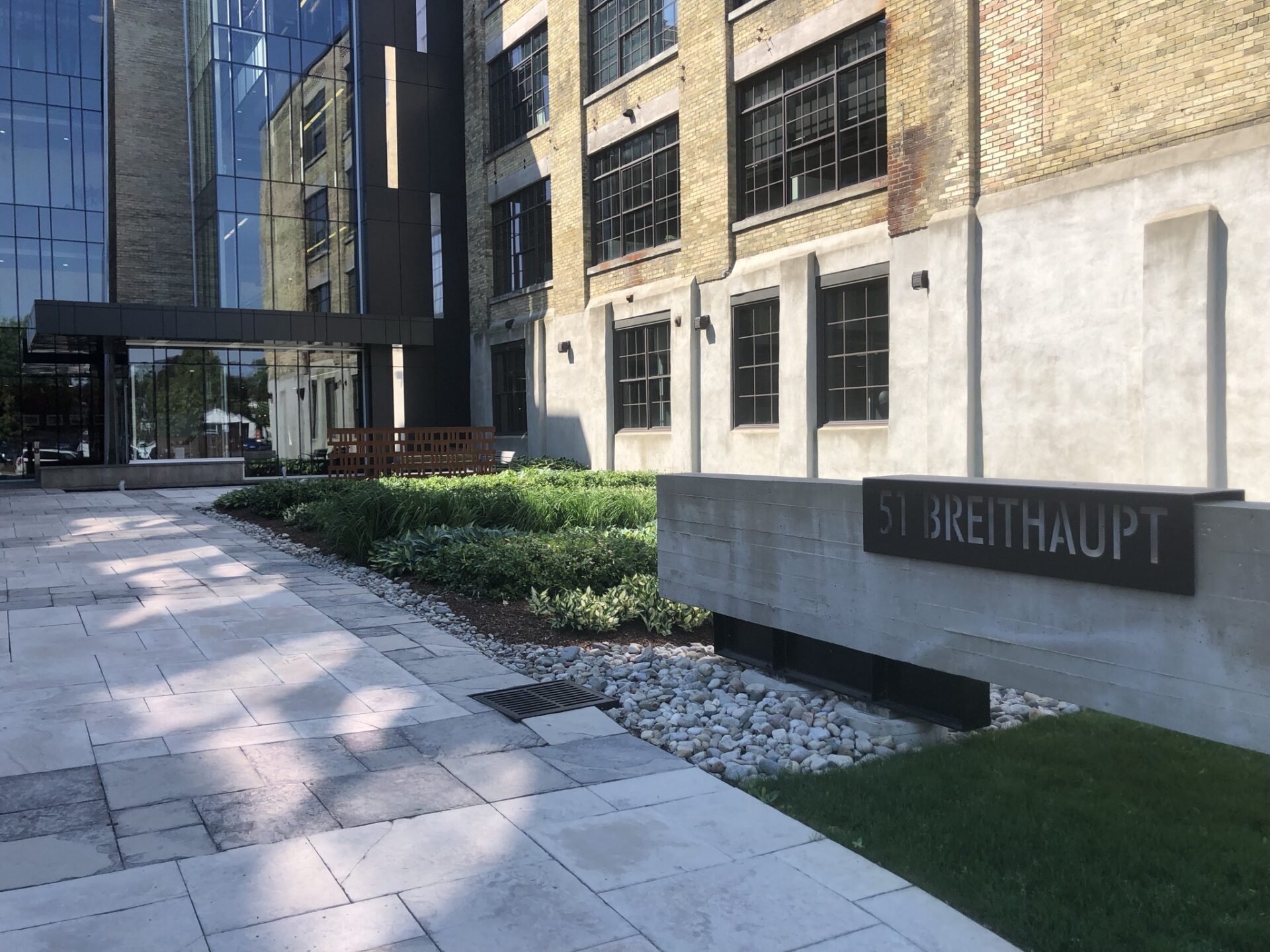 Modern office building with glass facade, brick walls, and a sign reading “51 Breithaupt.” Landscaped path with plants and rocks in the foreground.