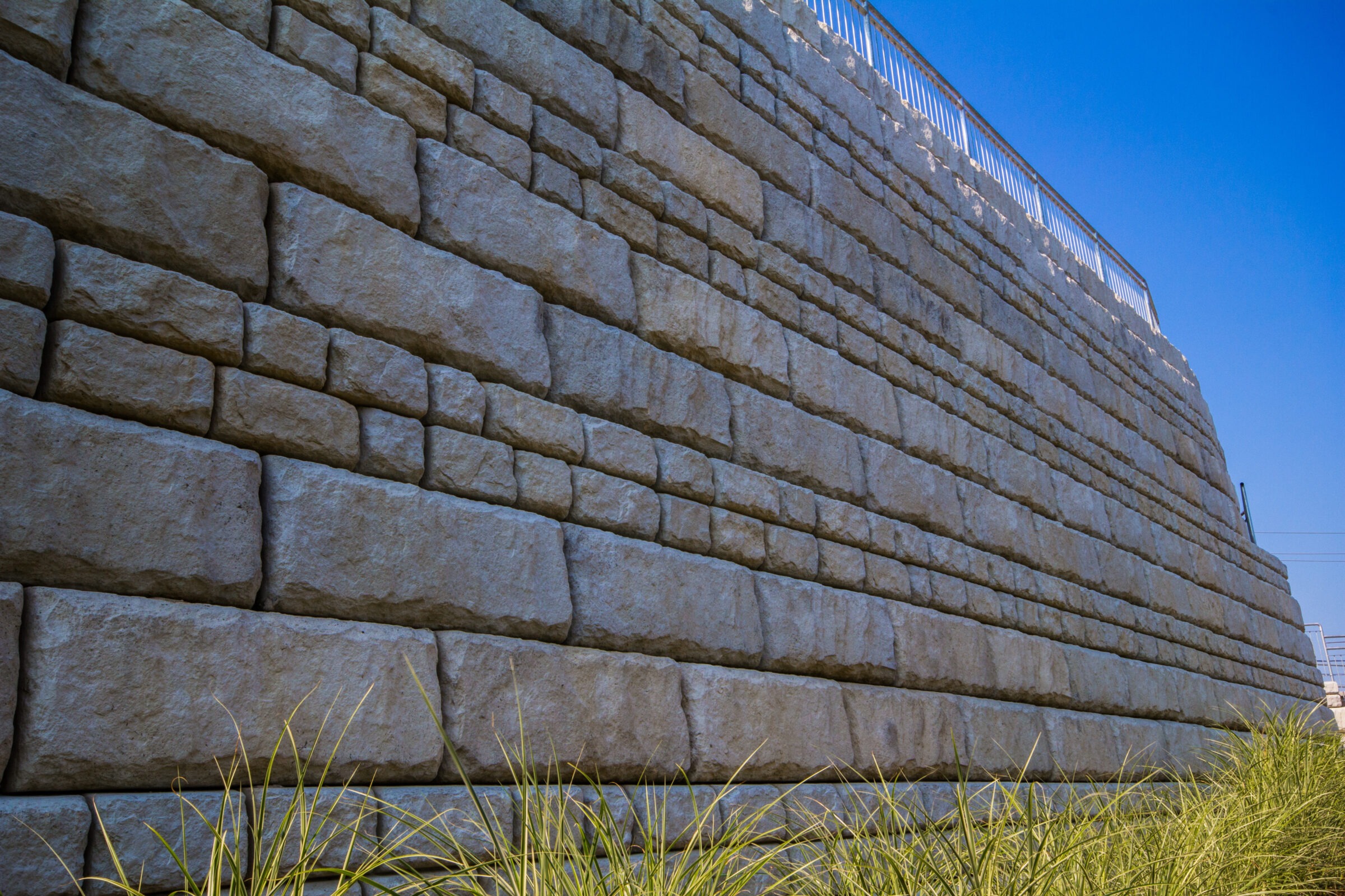 A sturdy stone retaining wall with a metal railing on top, framed by clear blue sky and green grass at the base.