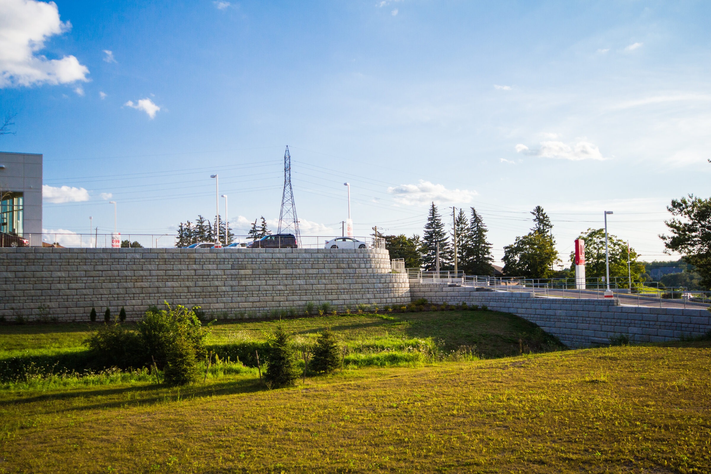 Sunny day with cars on a bridge, a stone wall, and green grass below. Blue sky with scattered clouds, trees nearby.