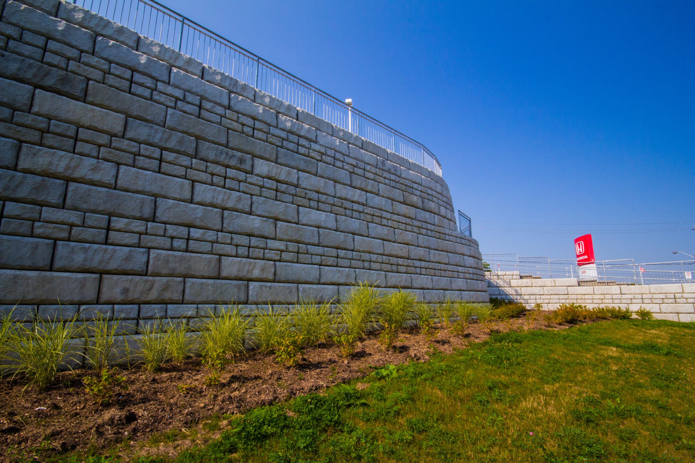 Stone retaining wall with metal railings, grassy slope with plants in foreground, clear blue sky, and a Honda sign in the background.