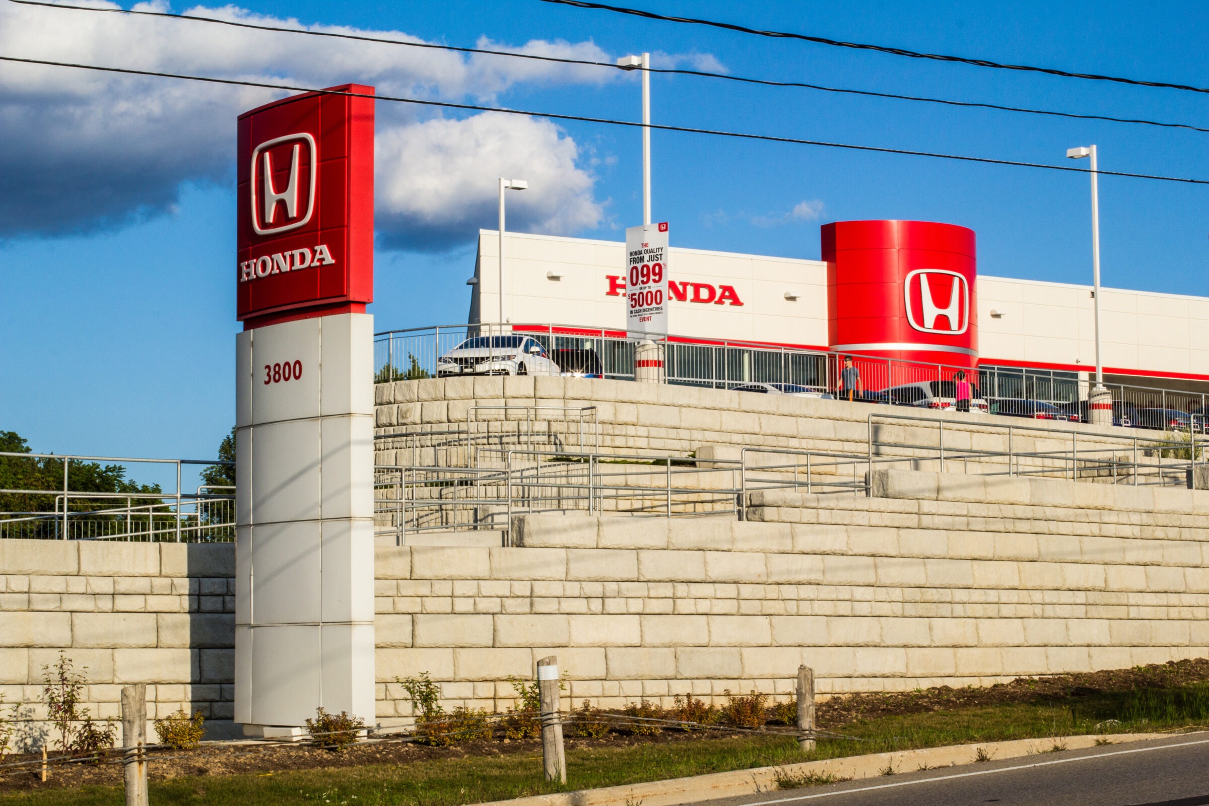 A Honda dealership with a red sign stands on a grassy hill, under a clear blue sky. Two people walk nearby.