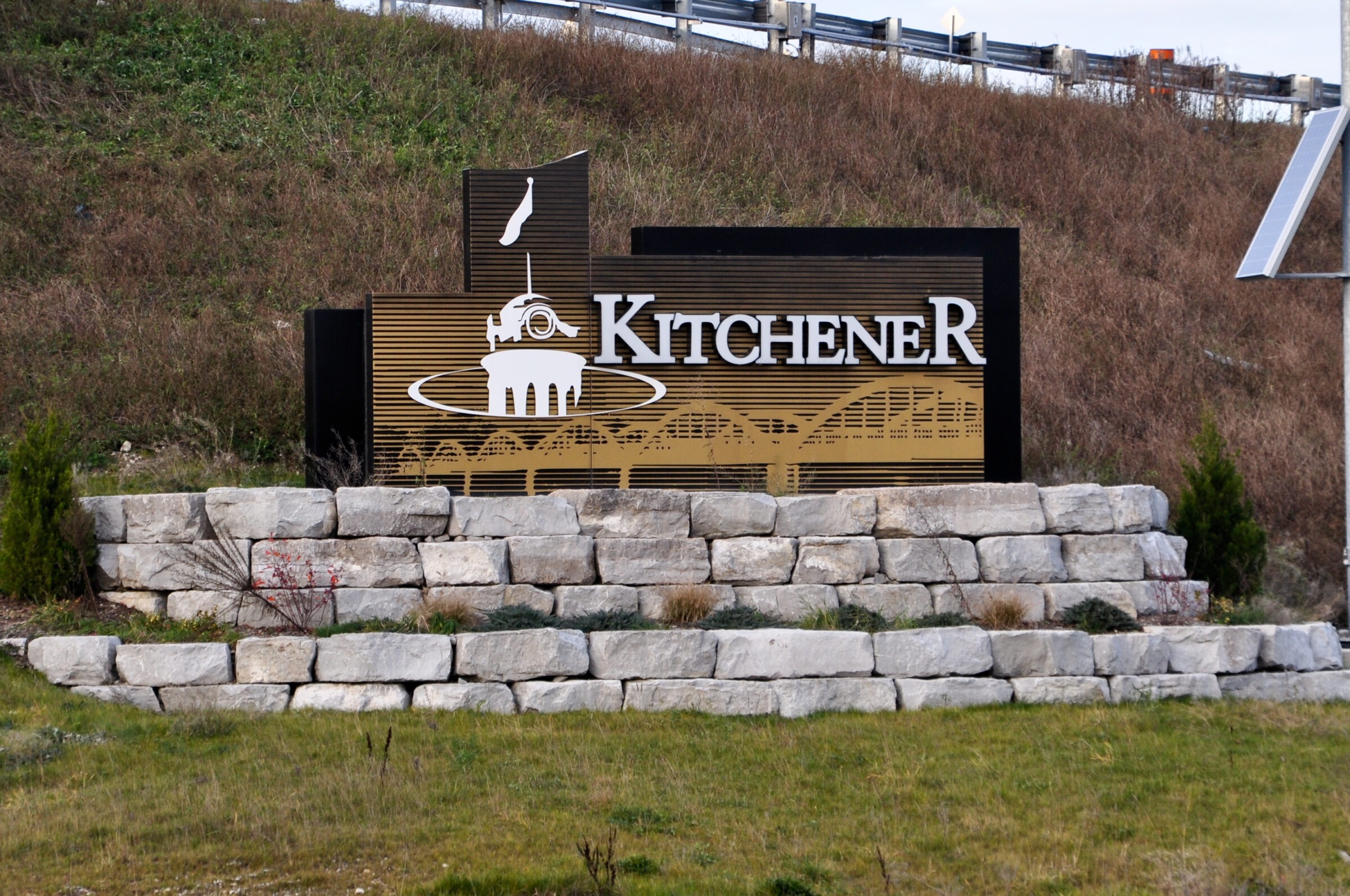 A sign with "Kitchener" and a city emblem atop stone landscaping, located by a grassy area and road barrier.