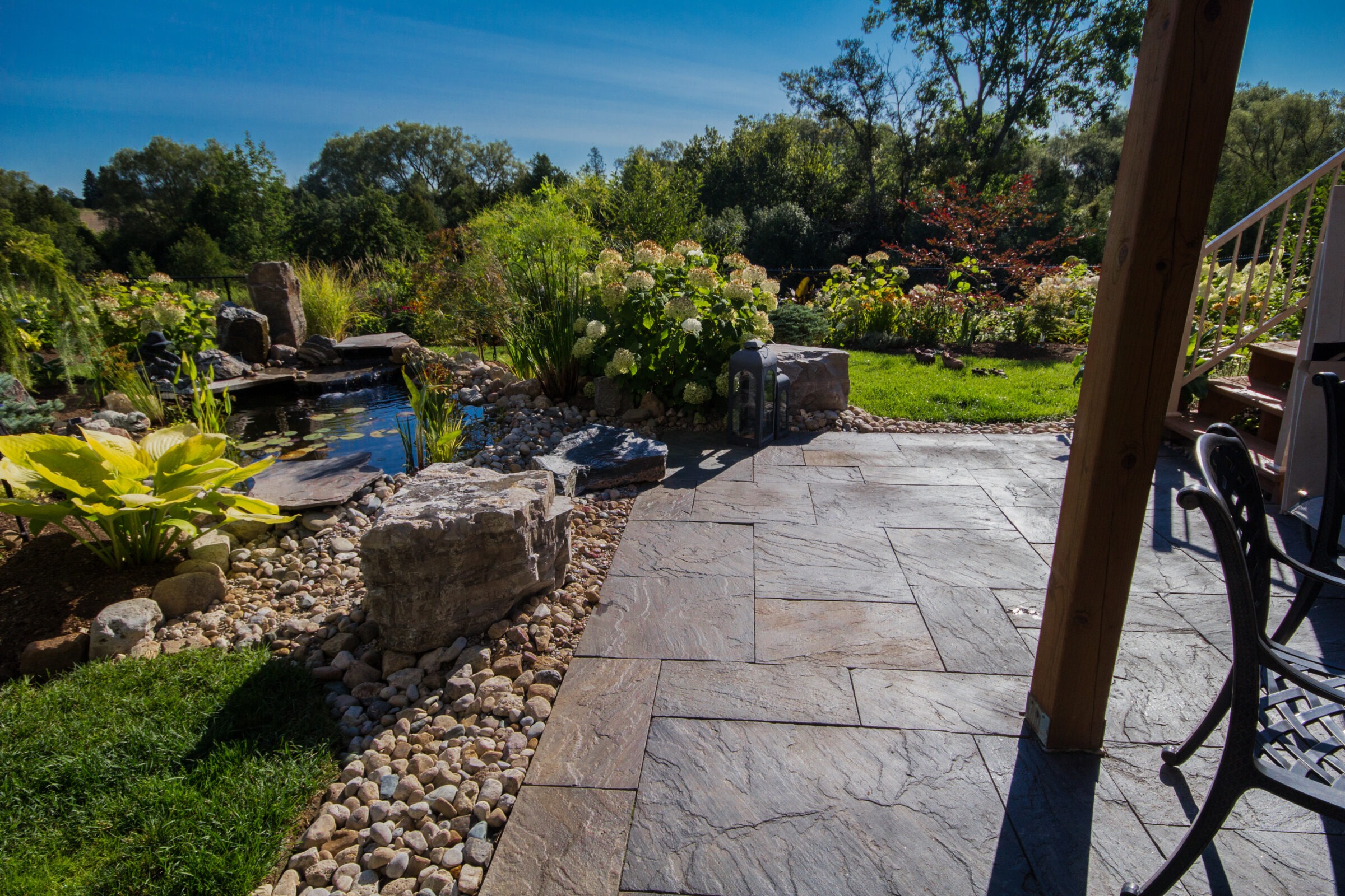 A serene garden setting features a stone patio, lush greenery, a tranquil pond, and chairs under a clear blue sky.