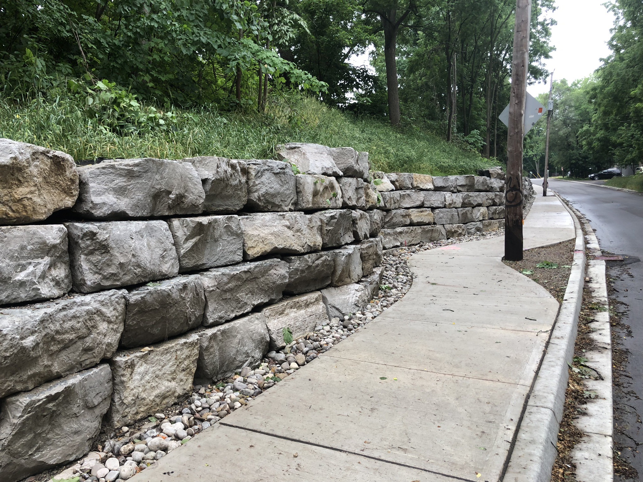 A winding sidewalk next to a sturdy stone retaining wall, flanked by lush greenery and a wooden utility pole, extends through a quiet street.