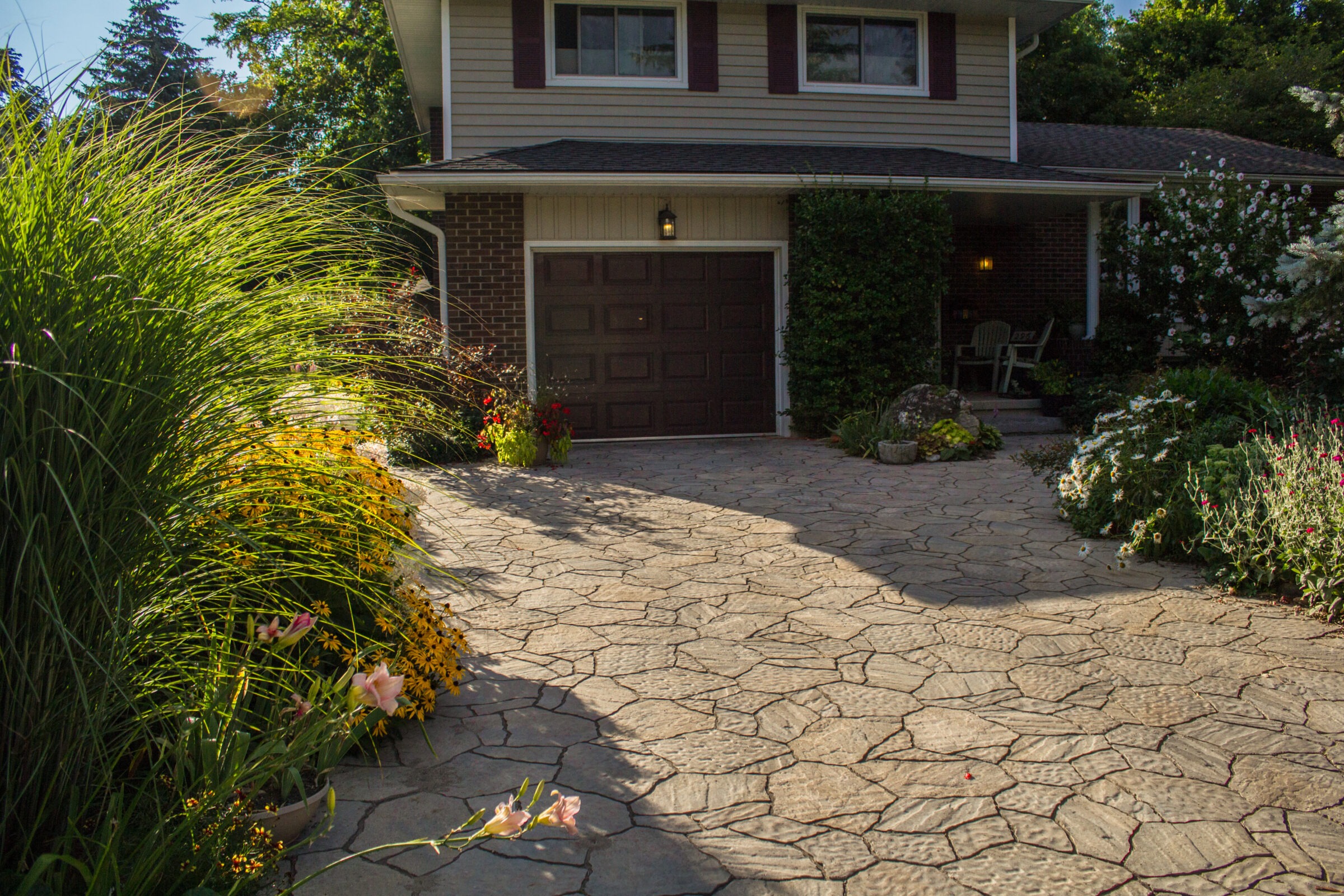 Suburban house with a stone driveway, surrounded by lush greenery and flowers. A cozy porch with a chair is partially visible.