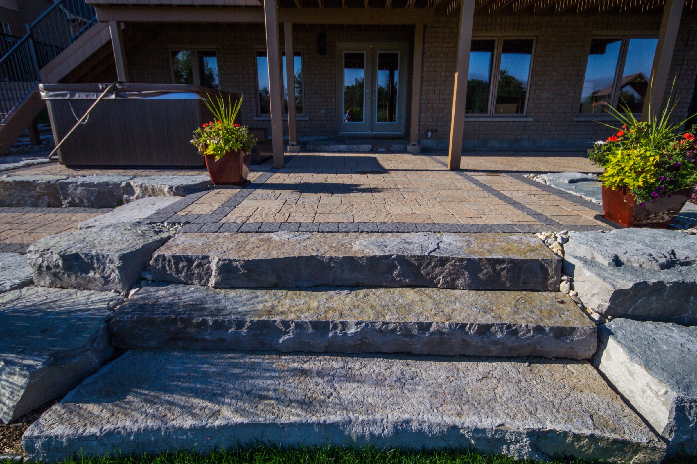 Stone steps lead to a patio with potted plants and a hot tub under a wooden pergola. Brick building facade visible.