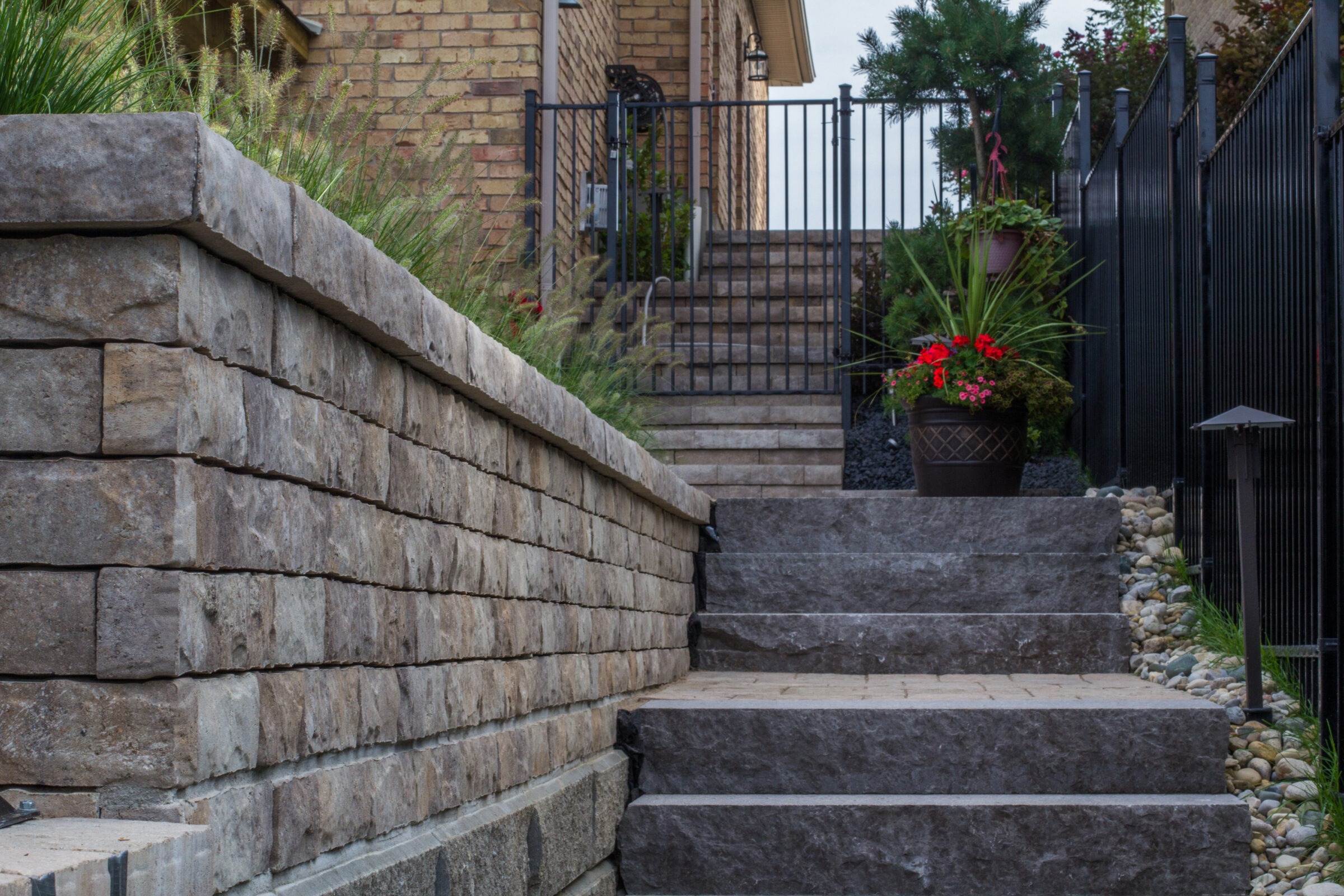 Stone steps lead up to a gated entrance, with potted plants and greenery lining the path, against a brick wall backdrop.