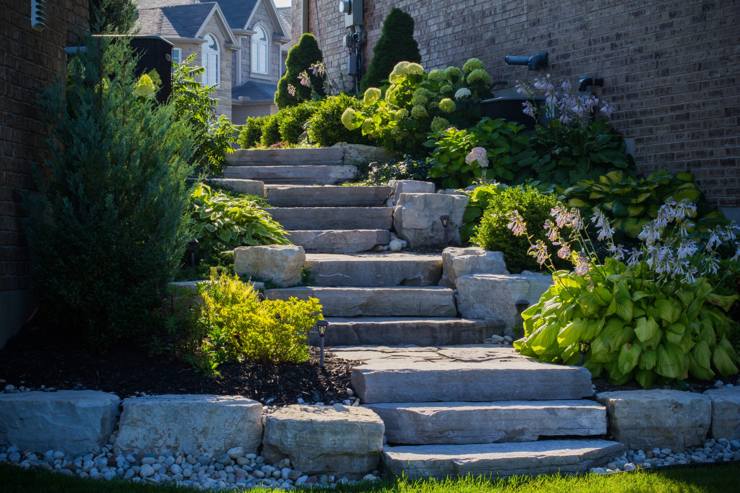 Stone steps lead through a lush garden with various plants, bordered by brick houses, creating a serene, landscaped outdoor setting.