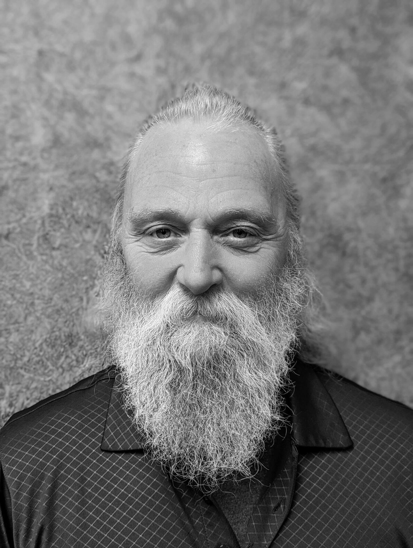 A person with a long beard and tied-back hair, wearing a patterned shirt, stands against a textured backdrop in this black-and-white portrait.
