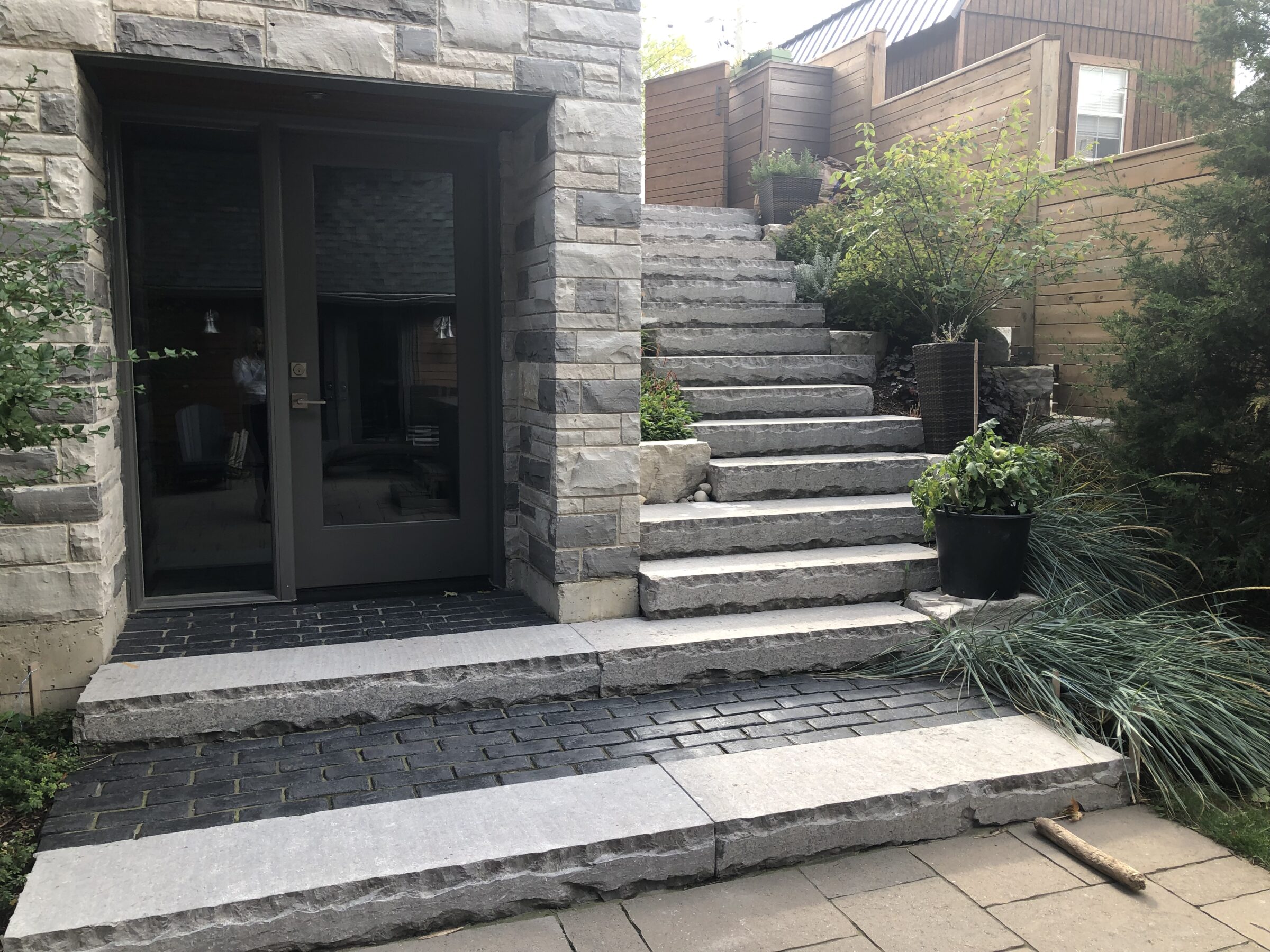 Stone steps leading to elevated patio with potted plants, bordered by wooden fencing, adjacent to glass door entrance and stone exterior wall.