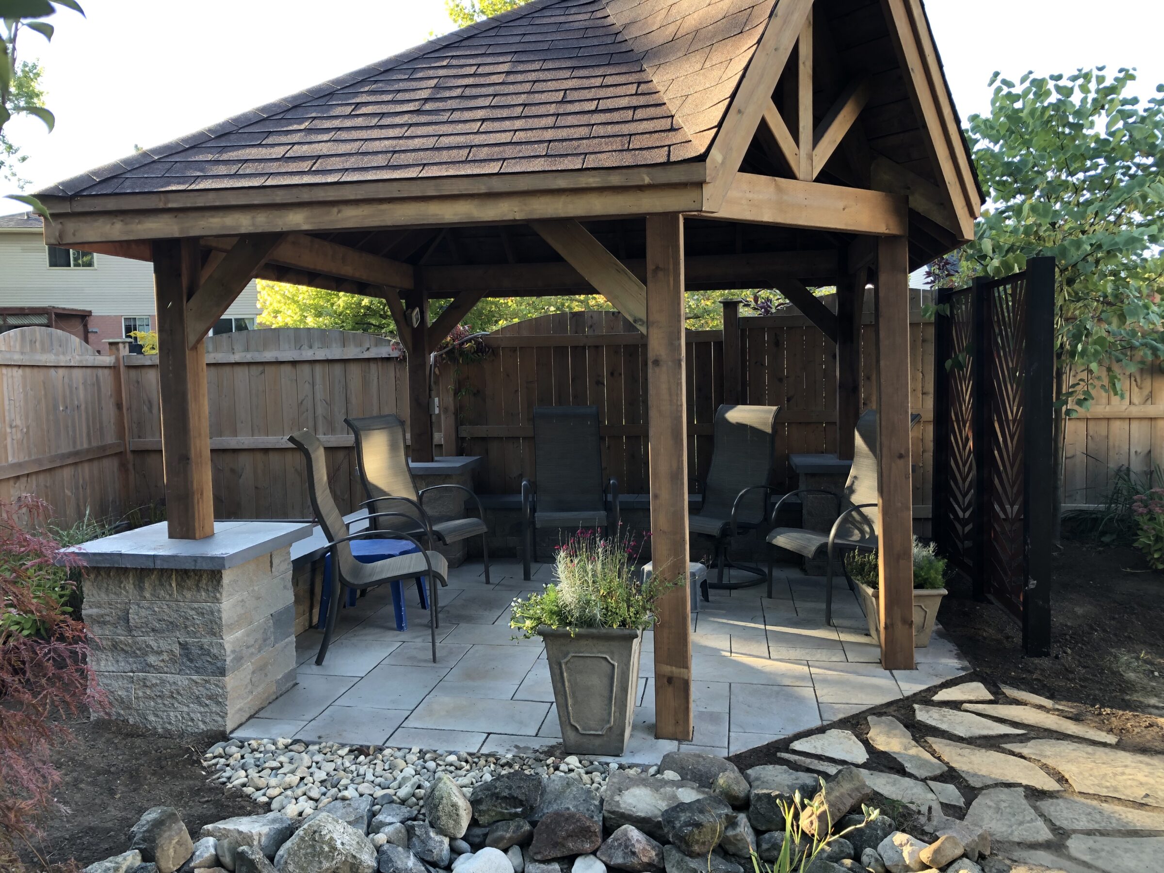A wooden gazebo with a tiled roof shelters several chairs on a stone patio, surrounded by a wooden fence and small garden.