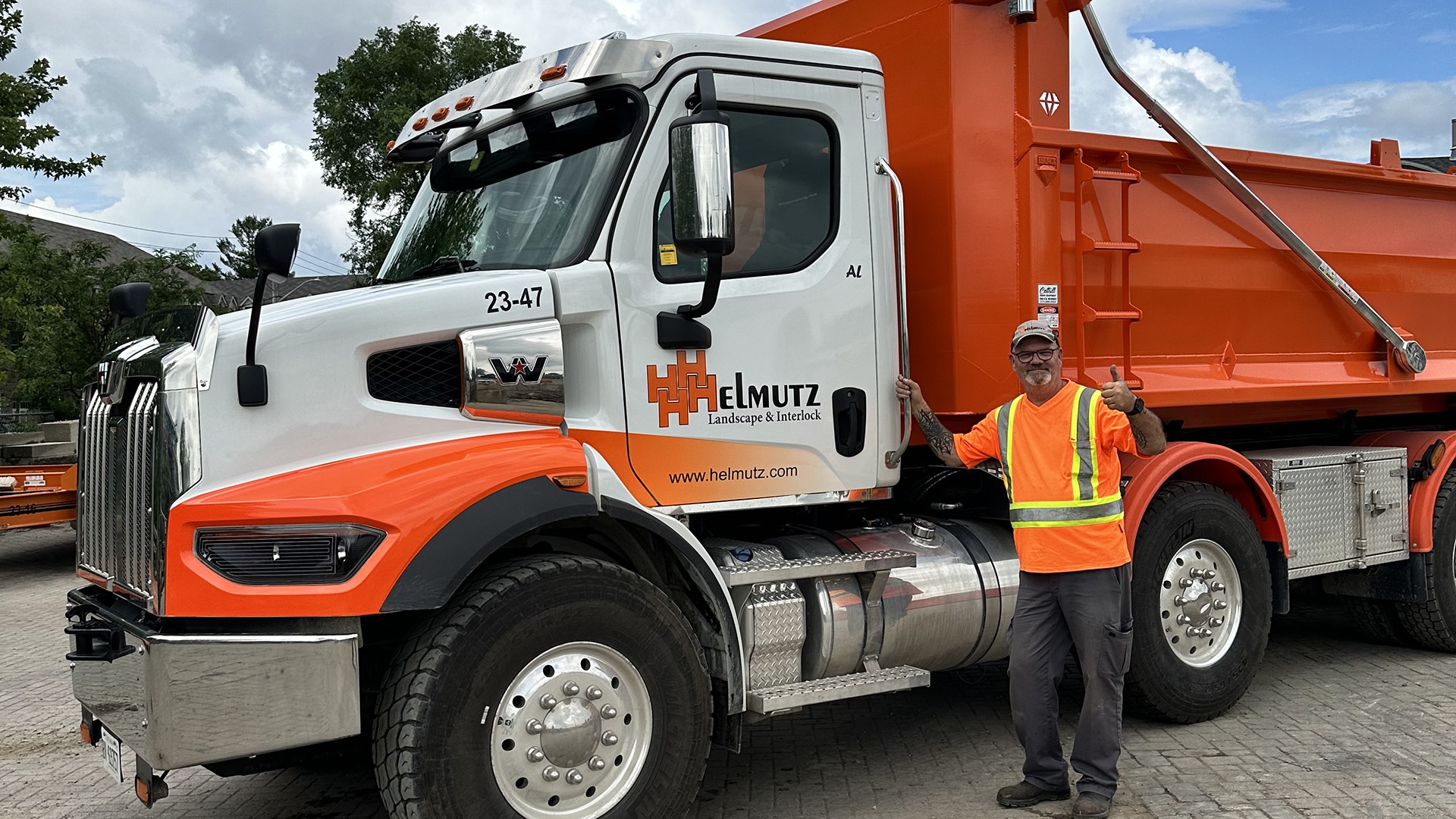 A person in a safety vest stands beside an orange-and-white Helmutz truck, giving a thumbs up. Overcast sky and trees in background.