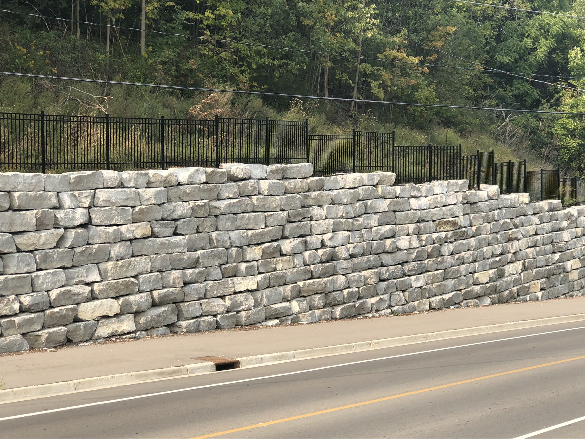 A stacked stone retaining wall lined with a black metal fence, bordered by a road and lush greenery in the background.