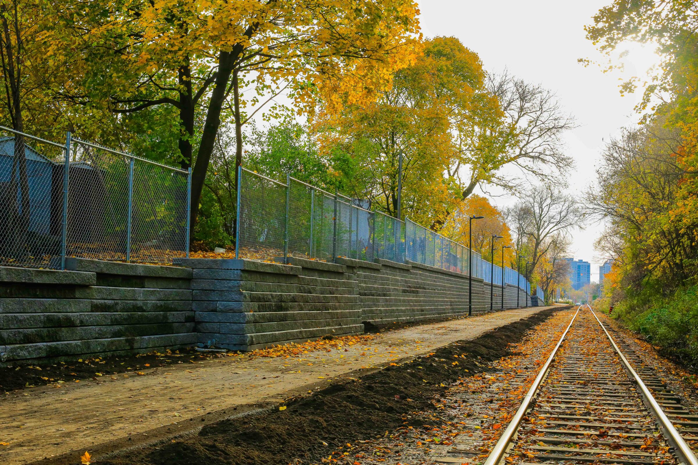 Railway tracks run through autumn trees beside a stone wall and fence, with a distant city skyline visible under a bright sky.