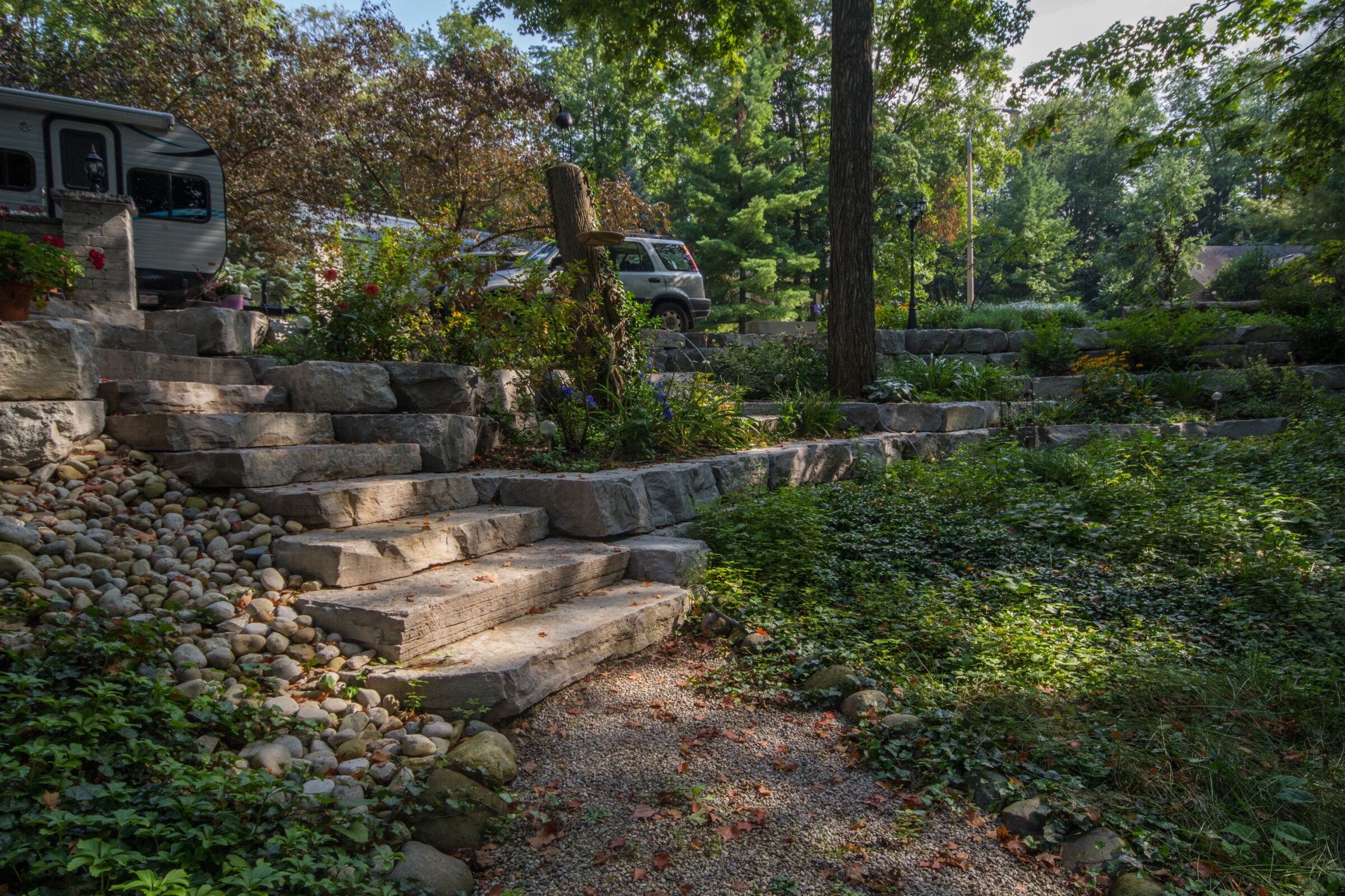Stone steps lead through a lush garden with trees and a pathway, accompanied by a parked SUV and trailer nearby.