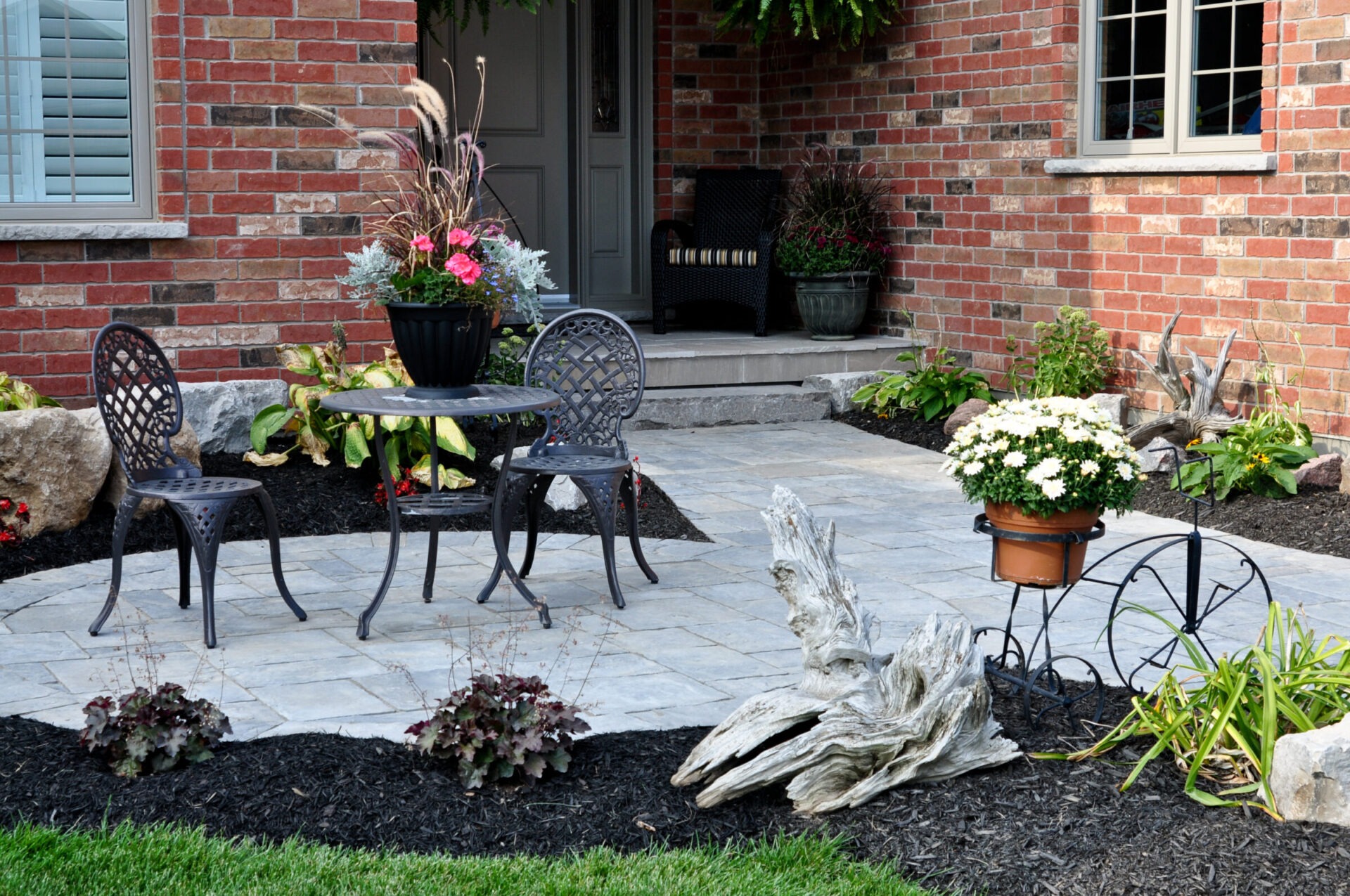 Front patio with stone pavers, black metal bistro set, flower planters, and landscaped garden beds.