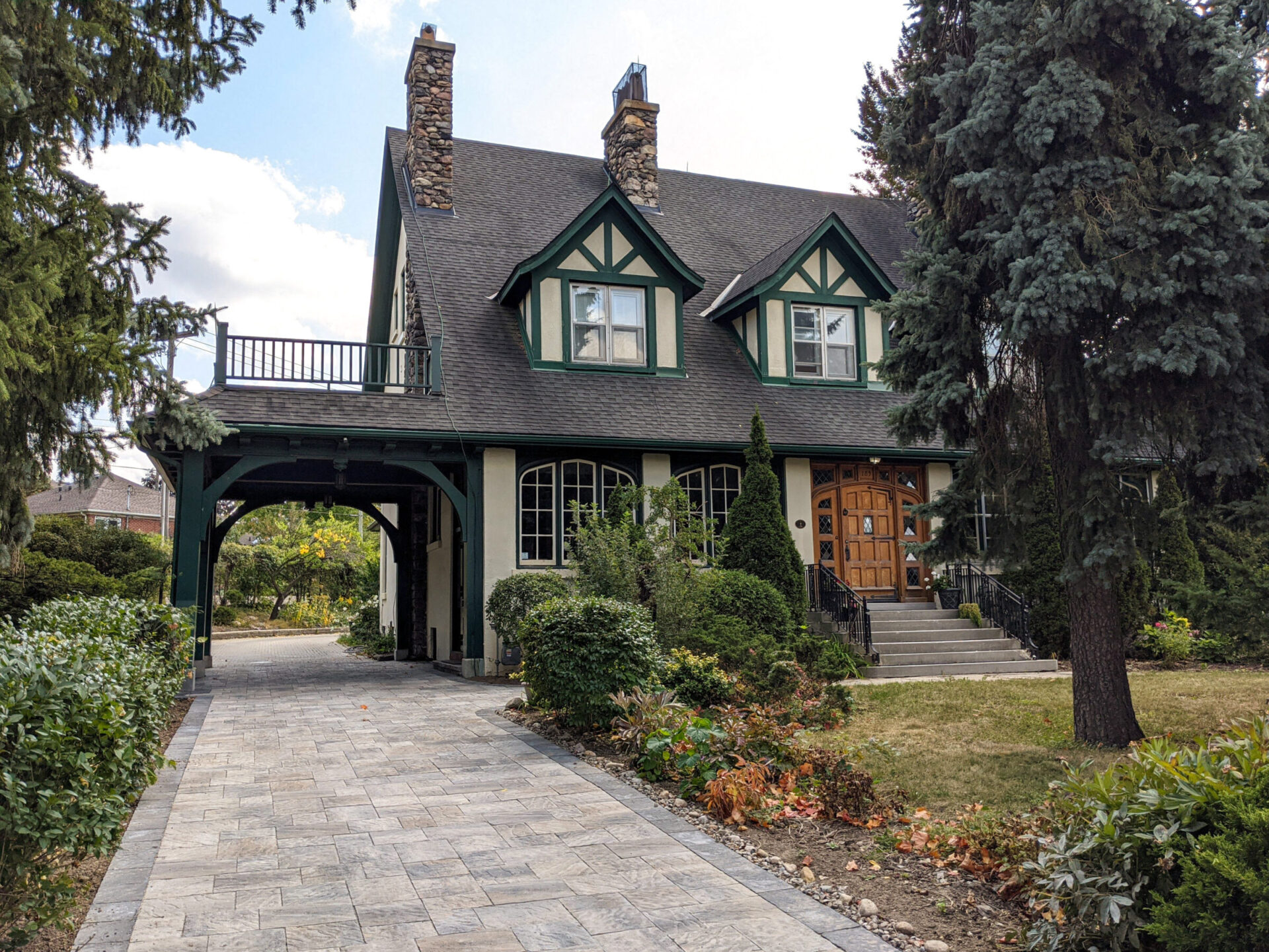 Charming Tudor-style house with stone chimneys and a paved driveway surrounded by mature trees.