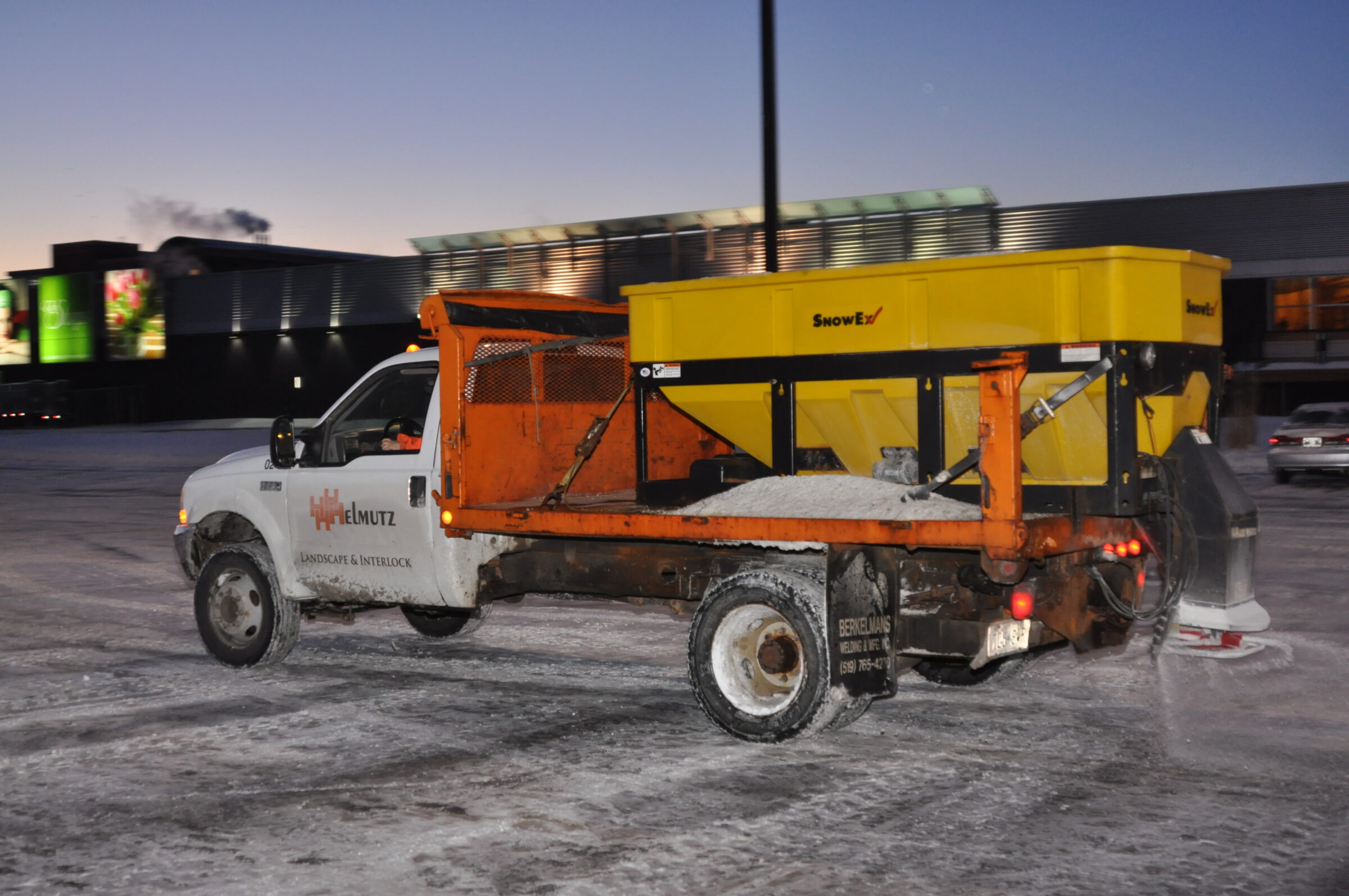 Truck with salt spreader clearing snow in parking lot at dusk