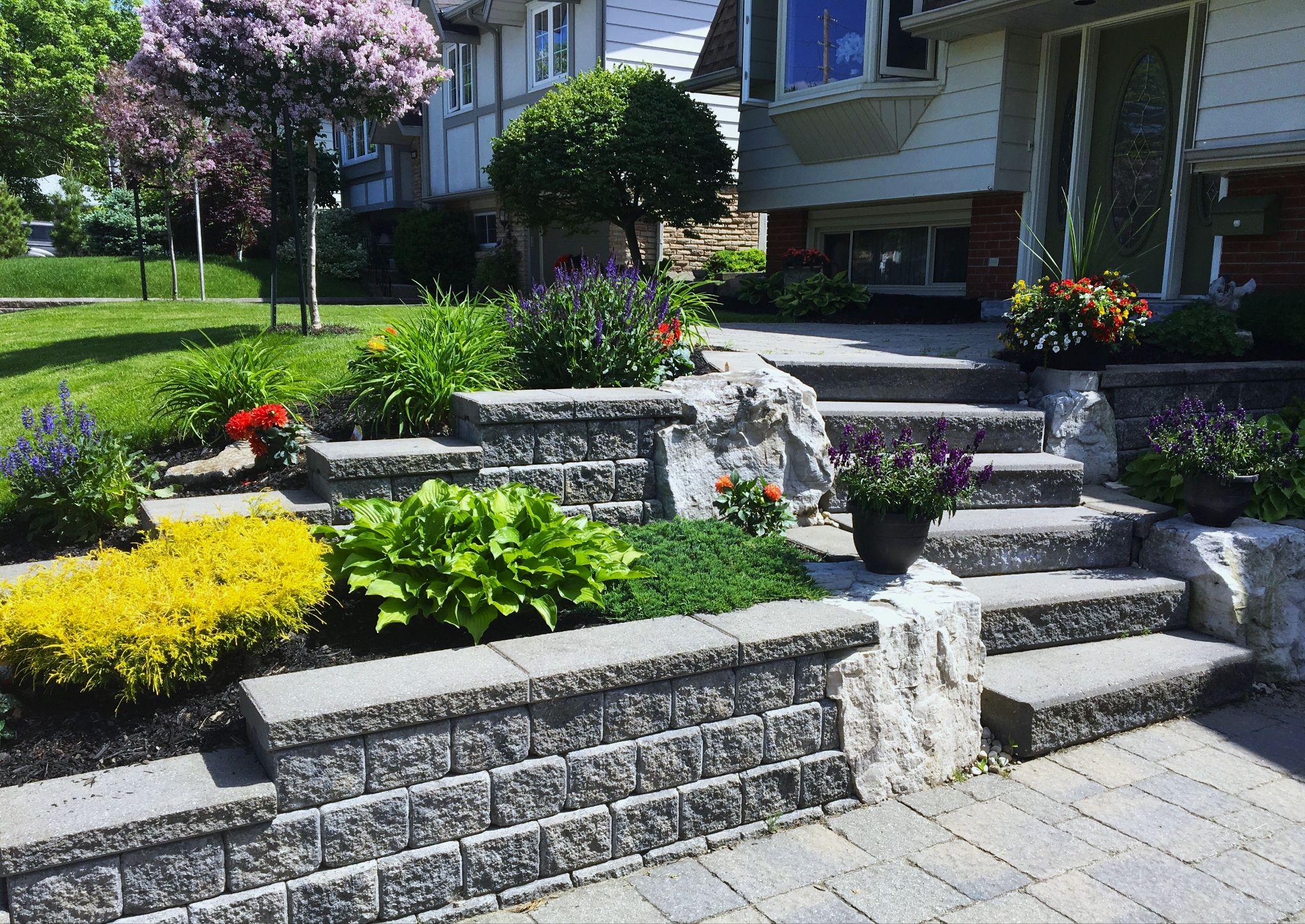 Tiered retaining wall with concrete blocks and natural stone accents framing front yard garden beds and steps.