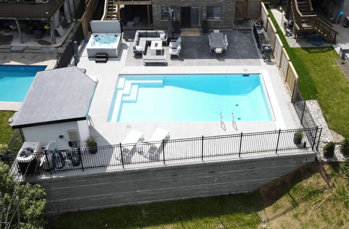 An aerial shot of a modern backyard pool, with a large, elegant retaining wall at the bottom of the image and the property, featuring a stone patio, seating area, and hot tub at the top of the image.